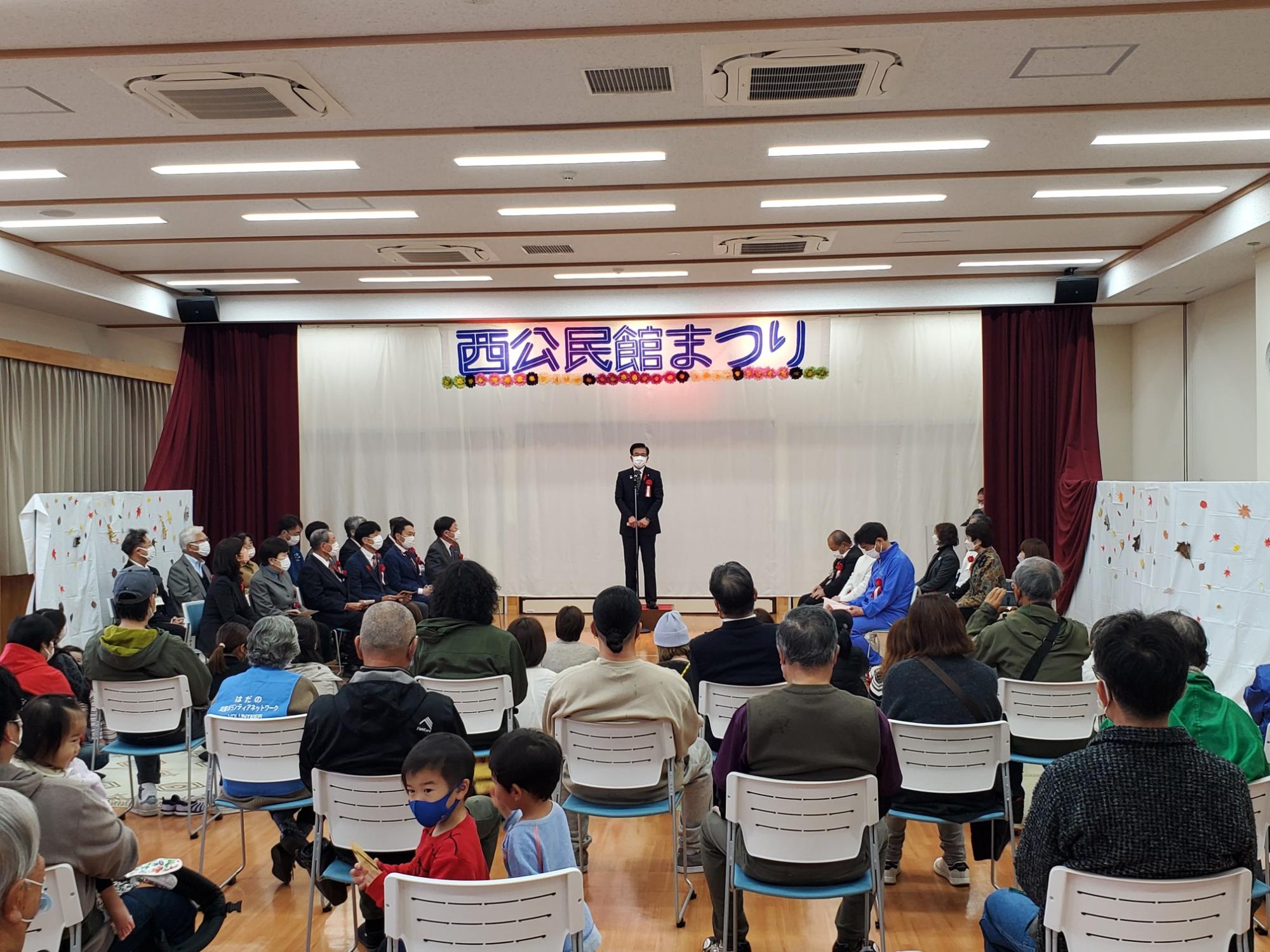 「西公民館まつり」と書かれた横断幕の舞台の中央でマスクを着用したスーツ姿の市長が、椅子に座った聴衆を前に話をしている写真