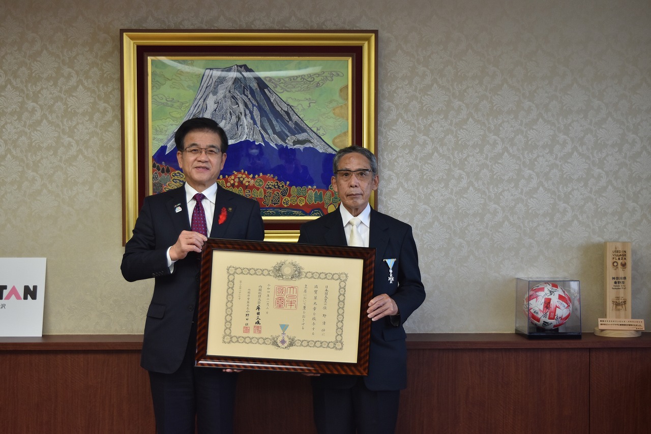 富士山の絵画の前に市長と勲章を胸につけた男性が額縁に入った賞状を持って並んで記念撮影をしている写真