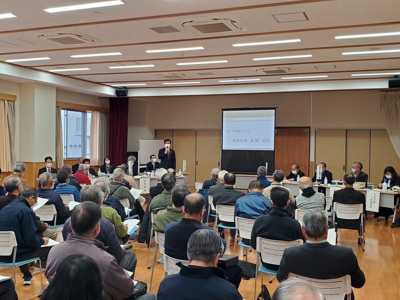 部屋の正面でスーツ姿の市長が、多くの人々が座っている会議室のプロジェクターの左側で話をしている西地区の市政懇談会の写真
