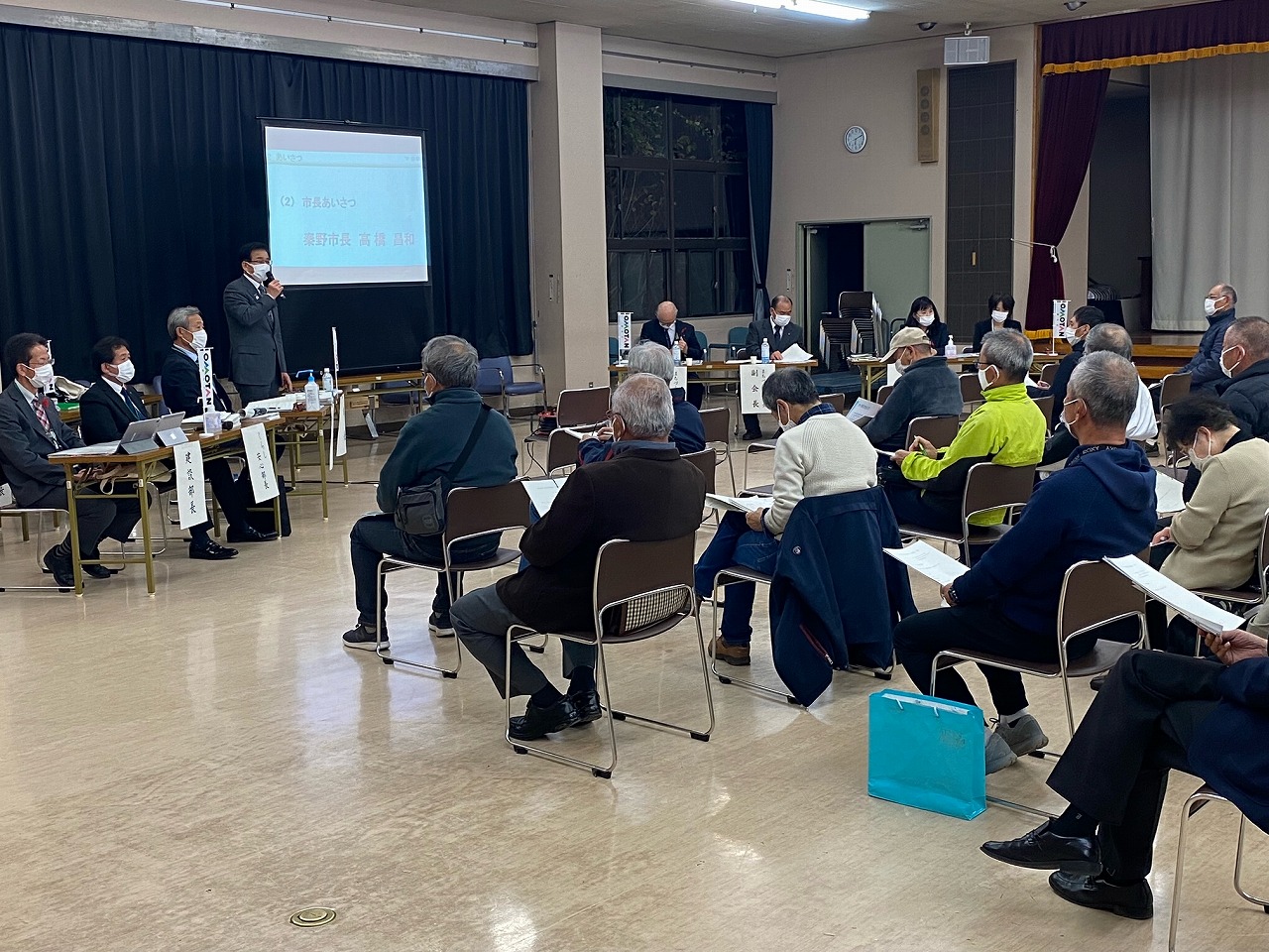 プロジェクターのスクリーンが設置された会場で、スーツ姿の市長がマイクを持ち話をしており、多数の参加者が椅子に座って聴講している大根地区の市政懇談会の写真