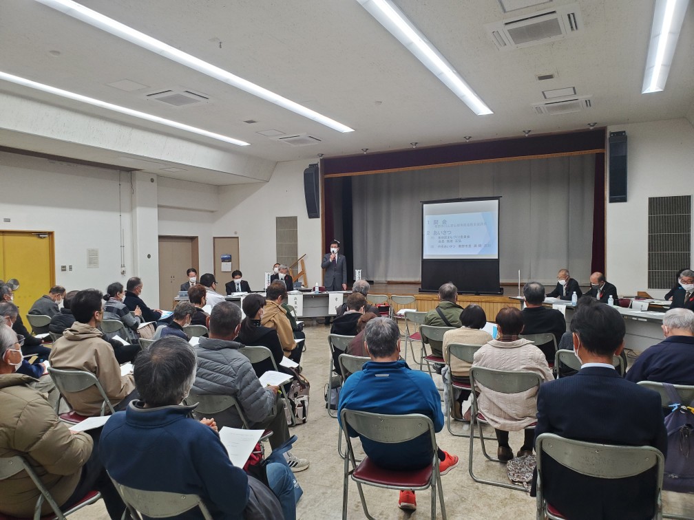 プロジェクターのスクリーンが設置された会場の前方で、マスクをスーツ姿の市長が、多数の参加者に向かってマイクで話している様子の東地区の市政懇談会の写真