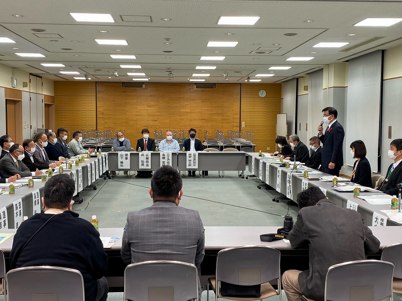 複数の人がコの字型に並んだテーブルに座り、立ってマイクを持って話すスーツ姿の市長の話を聞いている写真