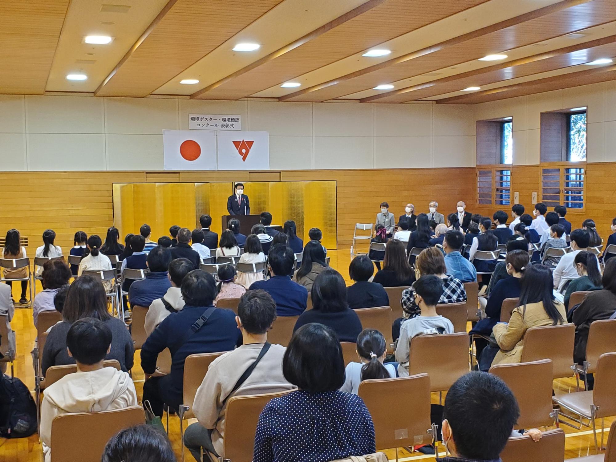 広い室内で、日本国旗と市の旗が掲げられた金屏風の前に立つ市長が、整然と並べられた椅子に座る多くの聴衆を前にして話をしている写真