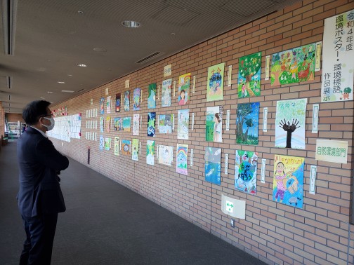 レンガの壁にたくさんの環境ポスターの作品が展示され、それをスーツ姿の市長が立ち止まってじっくり見ている様子の写真