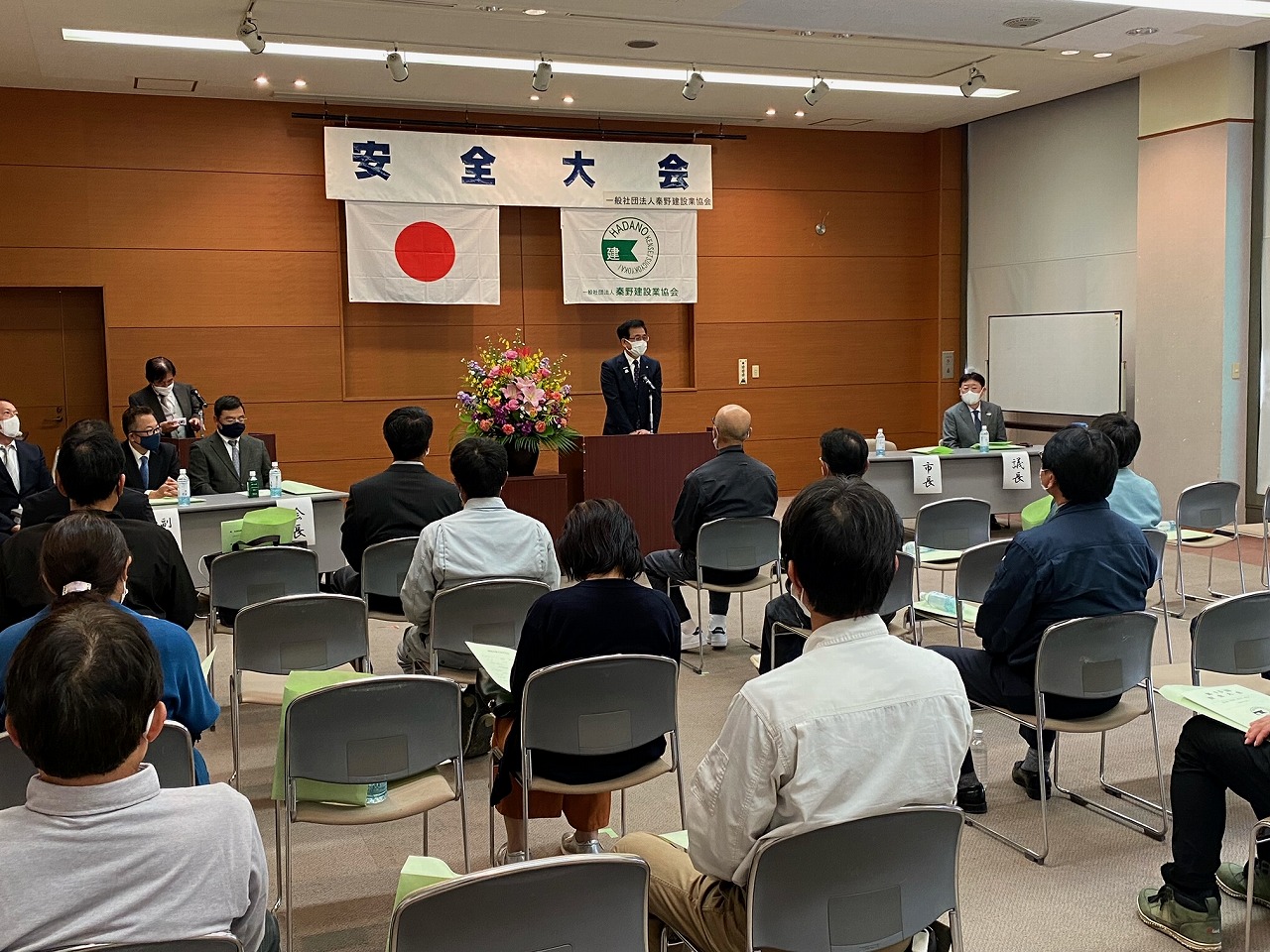 屋内で「安全大会」と書かれた横断幕と日の丸の旗が掲げられた壇上にスーツの市長が話をし、椅子に座った参加者が話を聞いている写真
