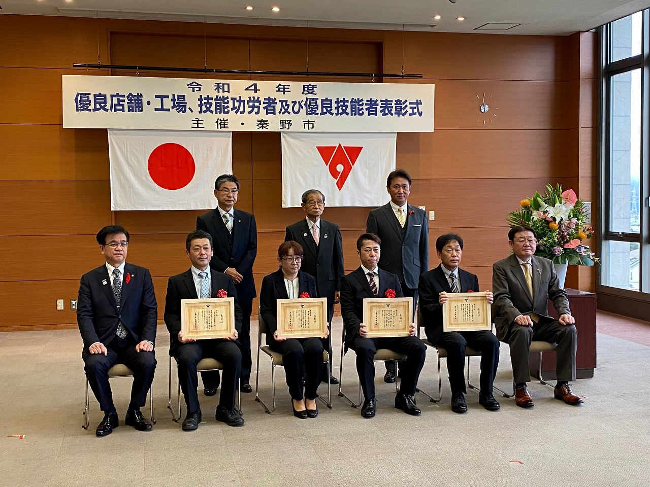 「令和4年度 優良店舗・工場・技能功労者及び優良技能者表彰式」と書かれた横断幕の前で、スーツ姿の市長と男女5人が椅子に座り4名が額に入った賞状を持ち後方に3名が立ち記念撮影をしている写真