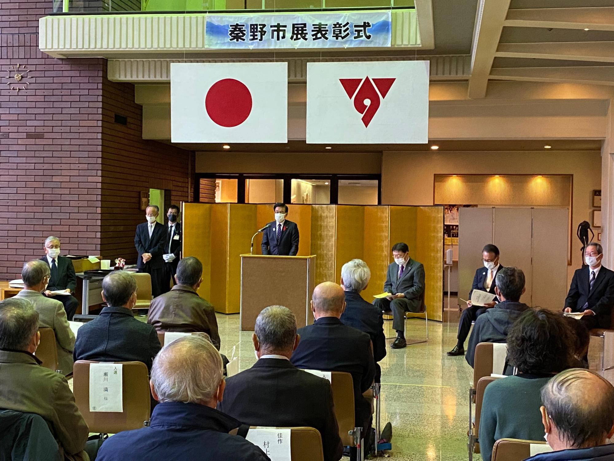 日本国旗と市の旗が掲げられた「秦野市展表彰式」の横断幕が吊式吊るさた金屏風の前で、スーツ姿の市長が、椅子に座った参加者たちに話をしている写真