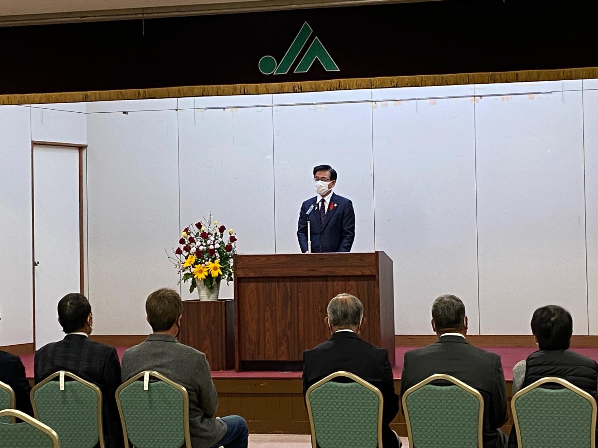 JAのロゴが掲げられた舞台で、花の飾られた演台からスーツ姿の市長が話をし、向かい合って関係者が椅子に座っている写真