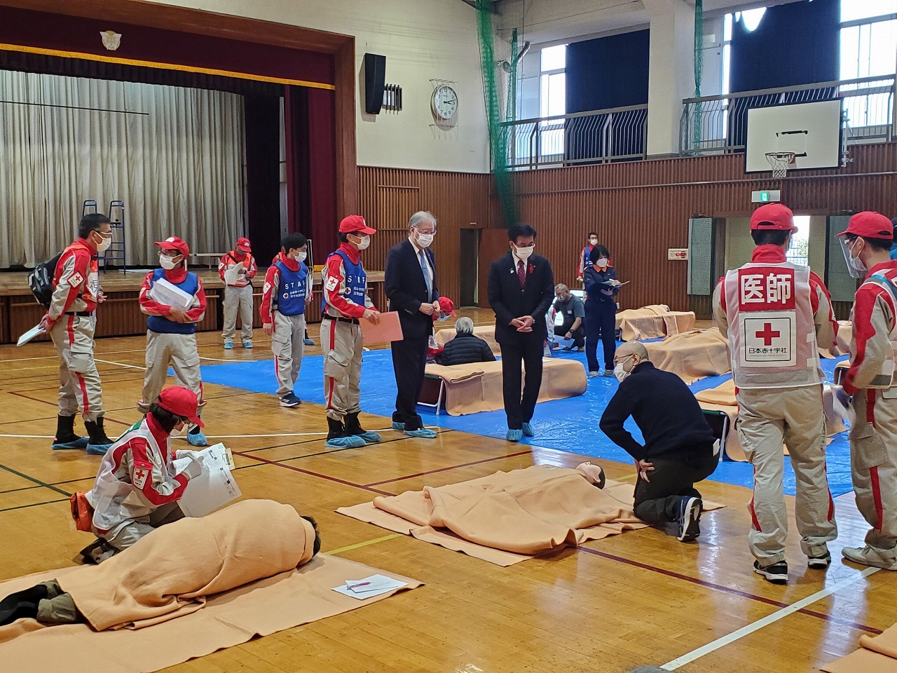 体育館での災害訓練で、毛布にくるまった人形や人を赤十字ボランティアの方々が医師、スタッフと書かれたビブスを着て訓練をしているのをスーツ姿の男性2人が視察している写真