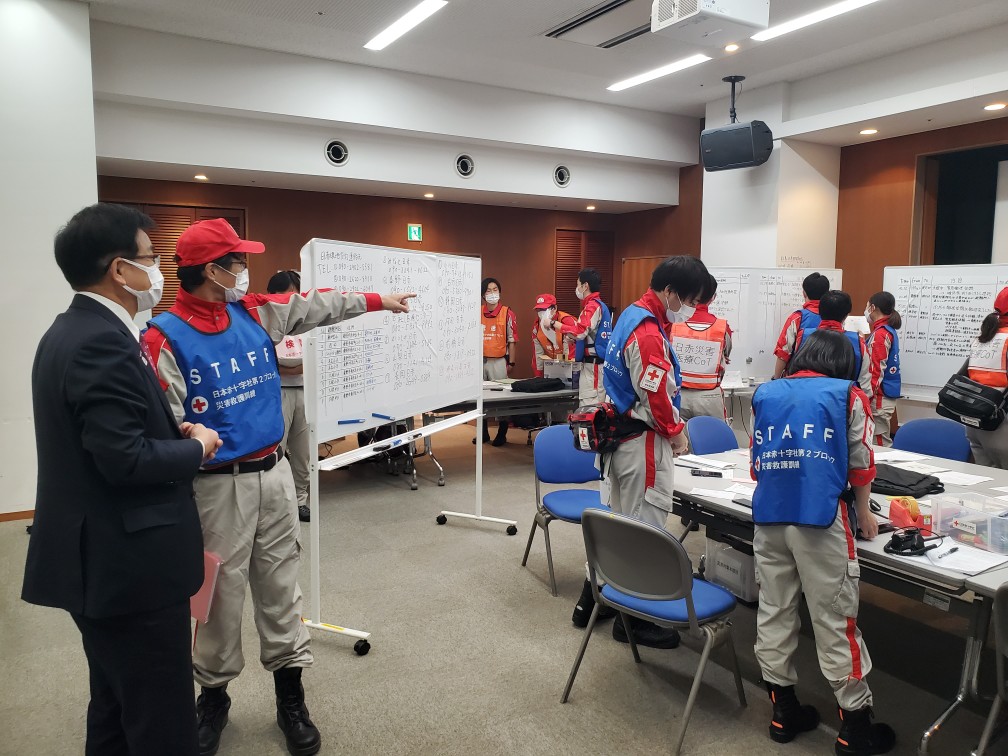 会議室でホワイトボードを使い、日本赤十字社のスタッフが被災時の対応などを確認しながら訓練を進めている様子をスーツ姿の市長が、赤いベストを着たスタッフから説明を受けている写真