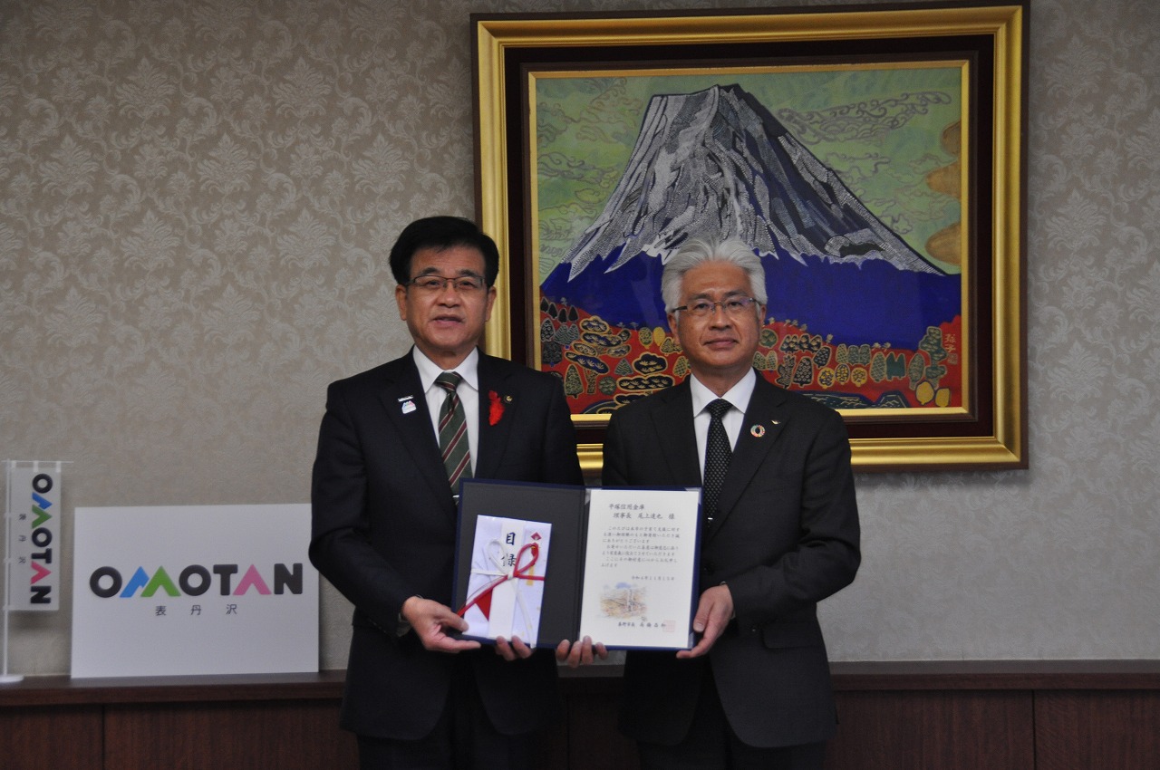 富士山の絵の前でスーツ姿の市長と尾上理事長が目録と賞状を持ち記念撮影をしている写真