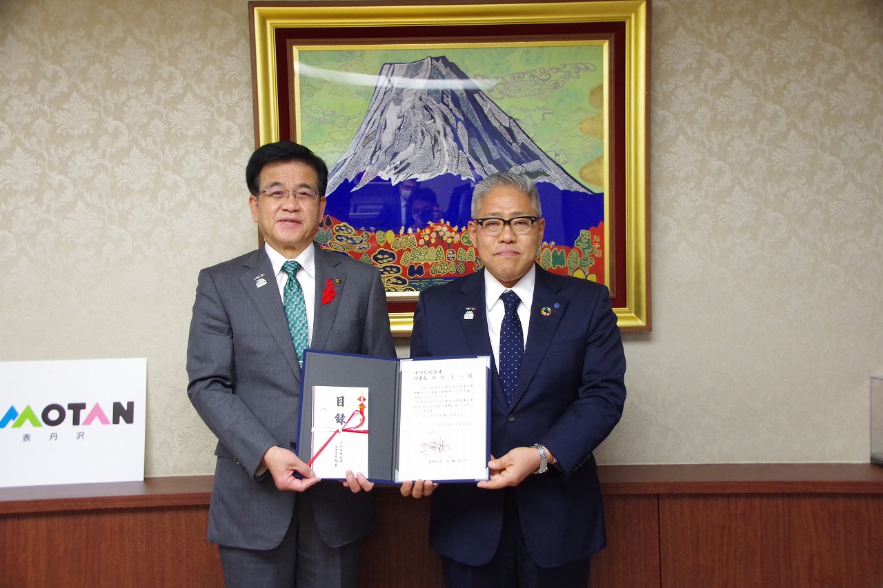 背景に富士山の絵画が飾られた部屋で、スーツ姿の市長と北村理事長が賞状と目録を両手で持ち記念撮影をしている写真
