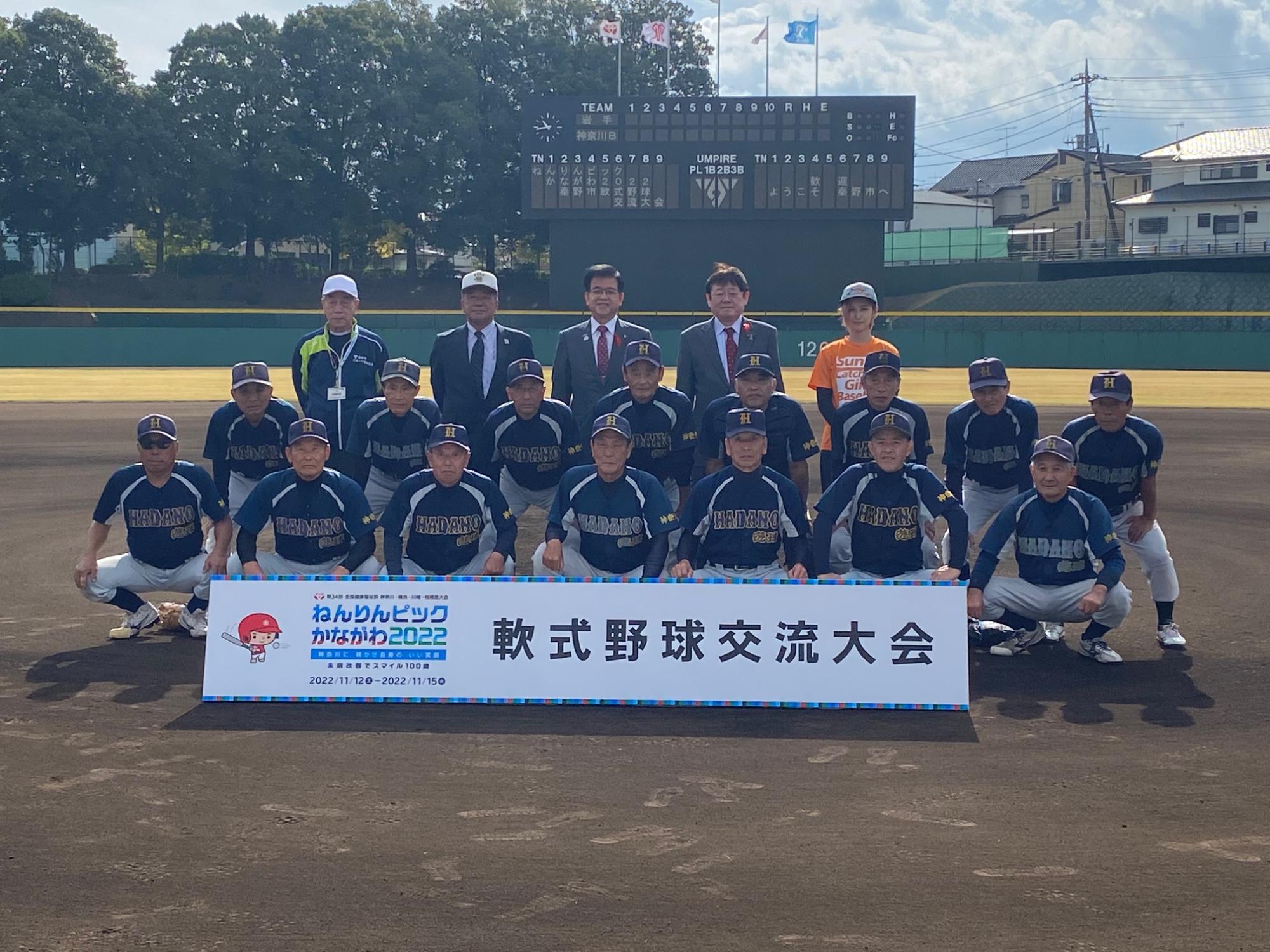 野球ユニフォームを着た選手たちが、野球場のグラウンドで「ねんりんピックかながわ2022 軟式野球交流大会」と書かれたボードの後ろで整列し、後列にはスーツ姿の関係者も並んで記念撮影している写真
