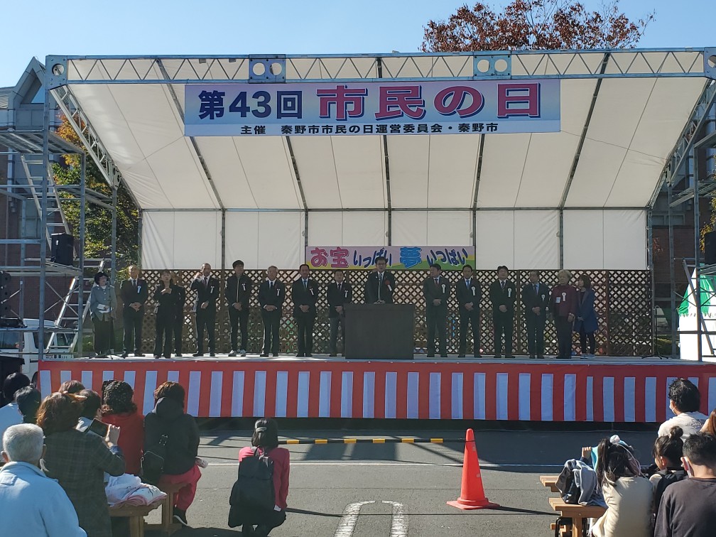 屋外の特設ステージ上でスーツ姿の複数の関係者が横一列に並び、スーツ姿の市長が舞台の中央に置かれた演壇に立ち話をしているのを大勢の観客がベンチに座って話を聞いている市民の日のイベントの写真