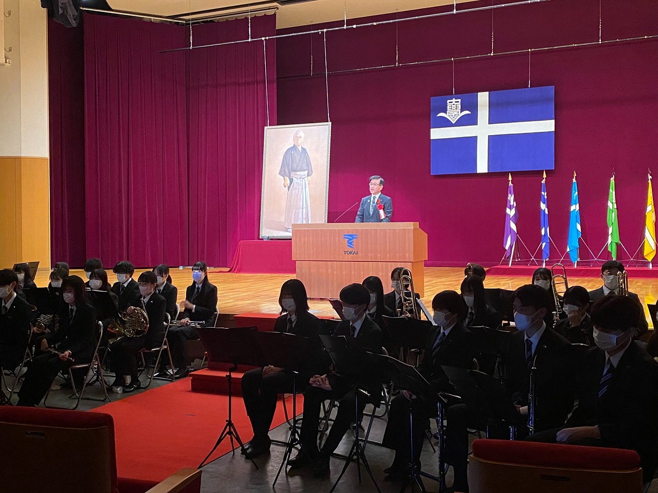 ステージに設けられた演台にスーツ姿の市長が話をし、舞台下には楽器を持った制服姿の学生たちが並んでいる写真