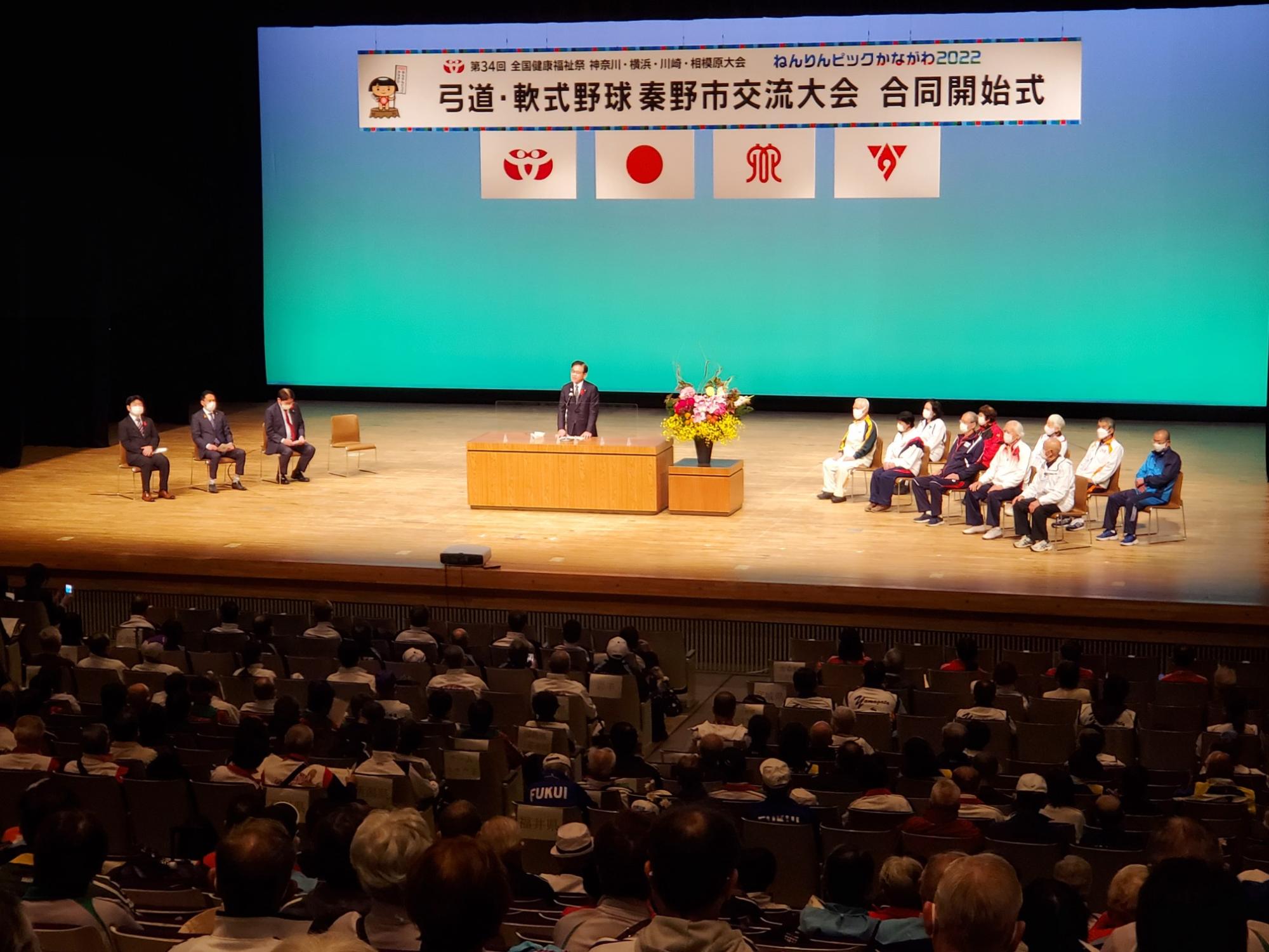 舞台上で「弓道・軟式野球秦野市交流大会合同開始式」と書かれた横断幕の下、スーツ姿の市長が演壇に立ち話をし、後方には出席者が座り、会場には複数の方が座っている写真