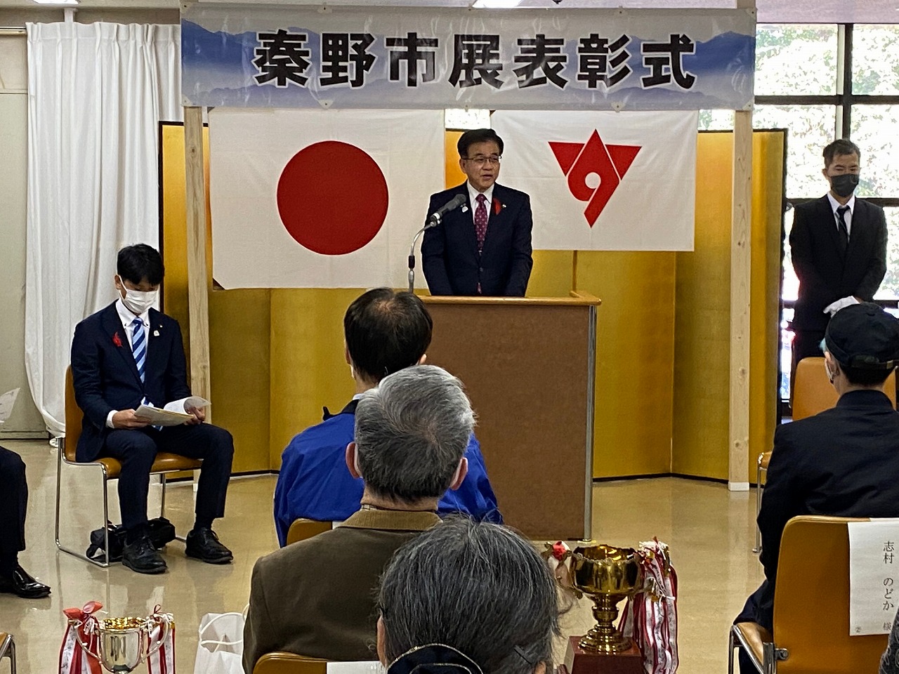 「令和4年度 保健衛生関係表彰式」と書かれた垂れ幕の下、舞台の上でスーツ姿の市長が女性に表彰状を渡しており、後方には日の丸と赤い市章の旗が掲げられている写真