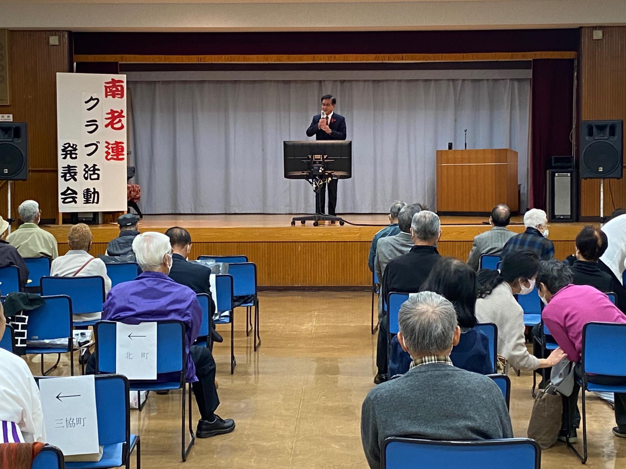 「南老連クラブ活動発表会」と書かれた看板が横に設置されている舞台の中央でスーツを着た市長が話をしているのを多くの年配者が椅子に座って話を聞いている写真