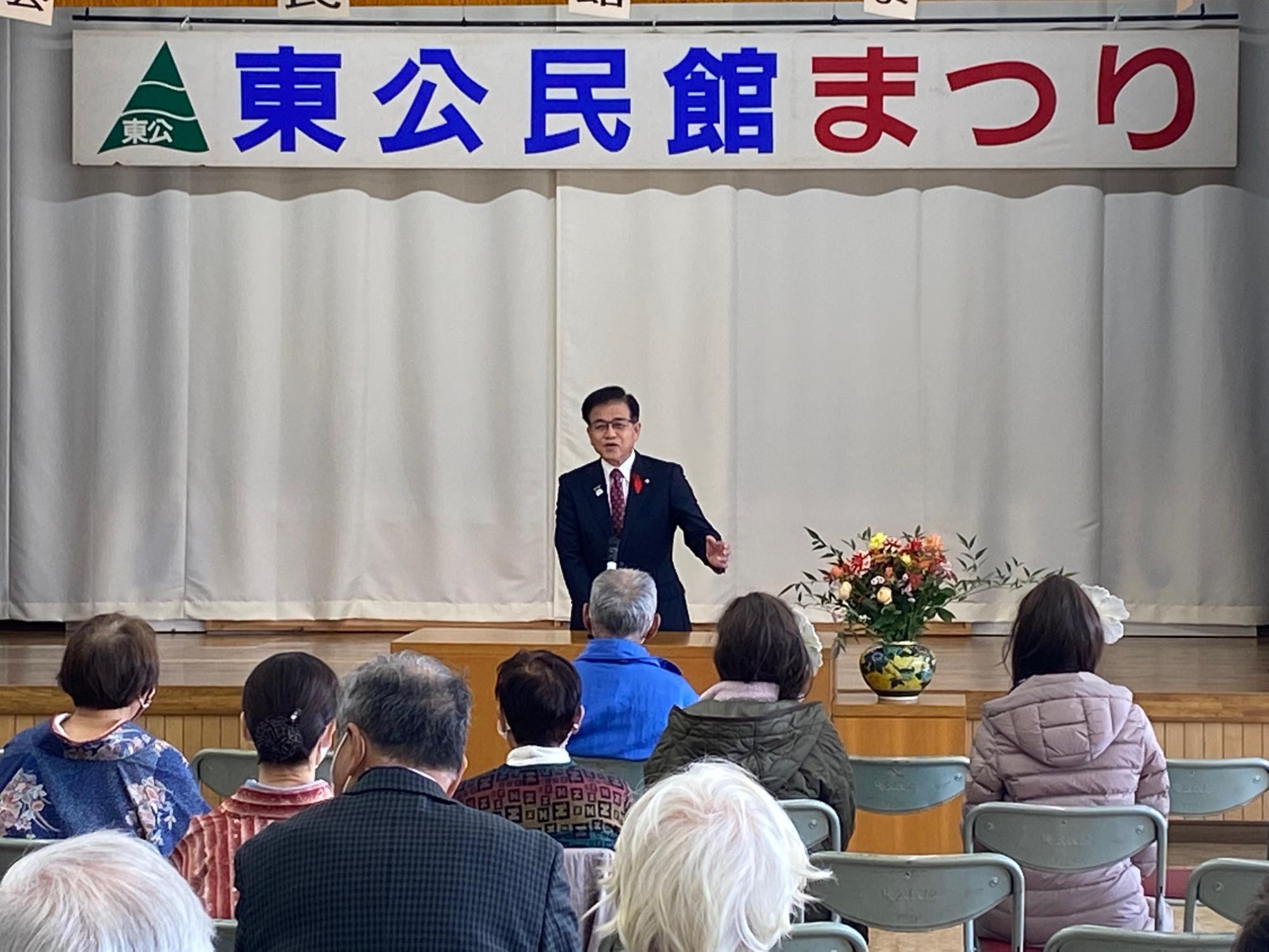 「東公民館まつり」と書かれた横断幕のある会場で、演台の横に花が飾られ、スーツ姿の市長がマイクを使って話をしている写真