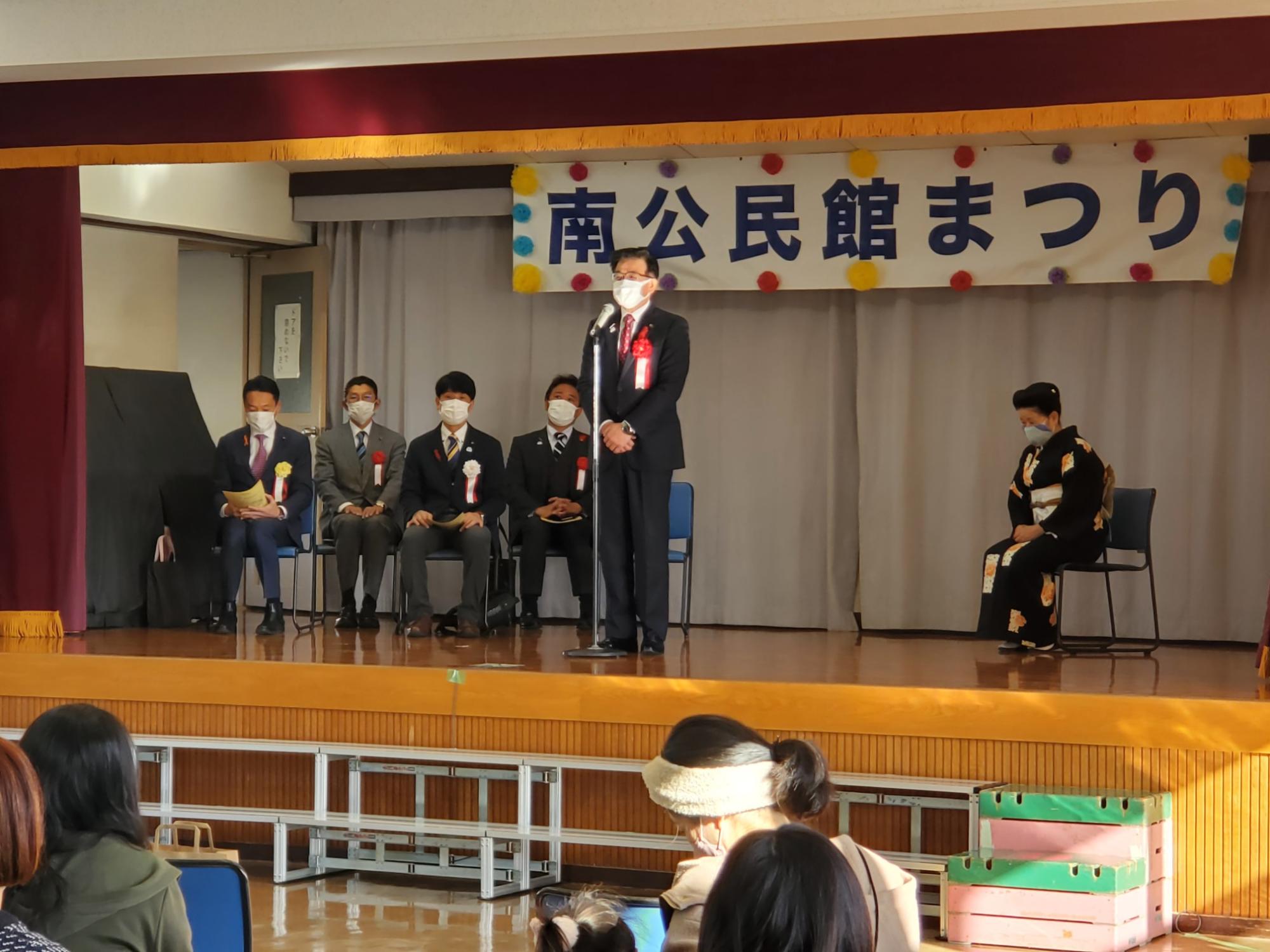 舞台上で、市長が中央におかれたマイクを持ってスピーチしており、その背景には「南公民館まつり」と書かれた横断幕が掲げられている写真