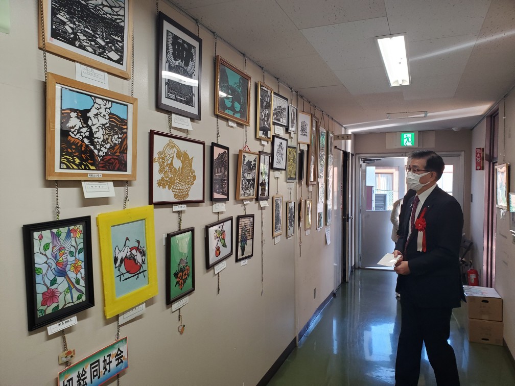 白い壁の長い廊下で、スーツ姿の男性がマスクを着用して立ち、壁には切り絵や版画と思われる数多くの額縁に入った作品が飾られている写真