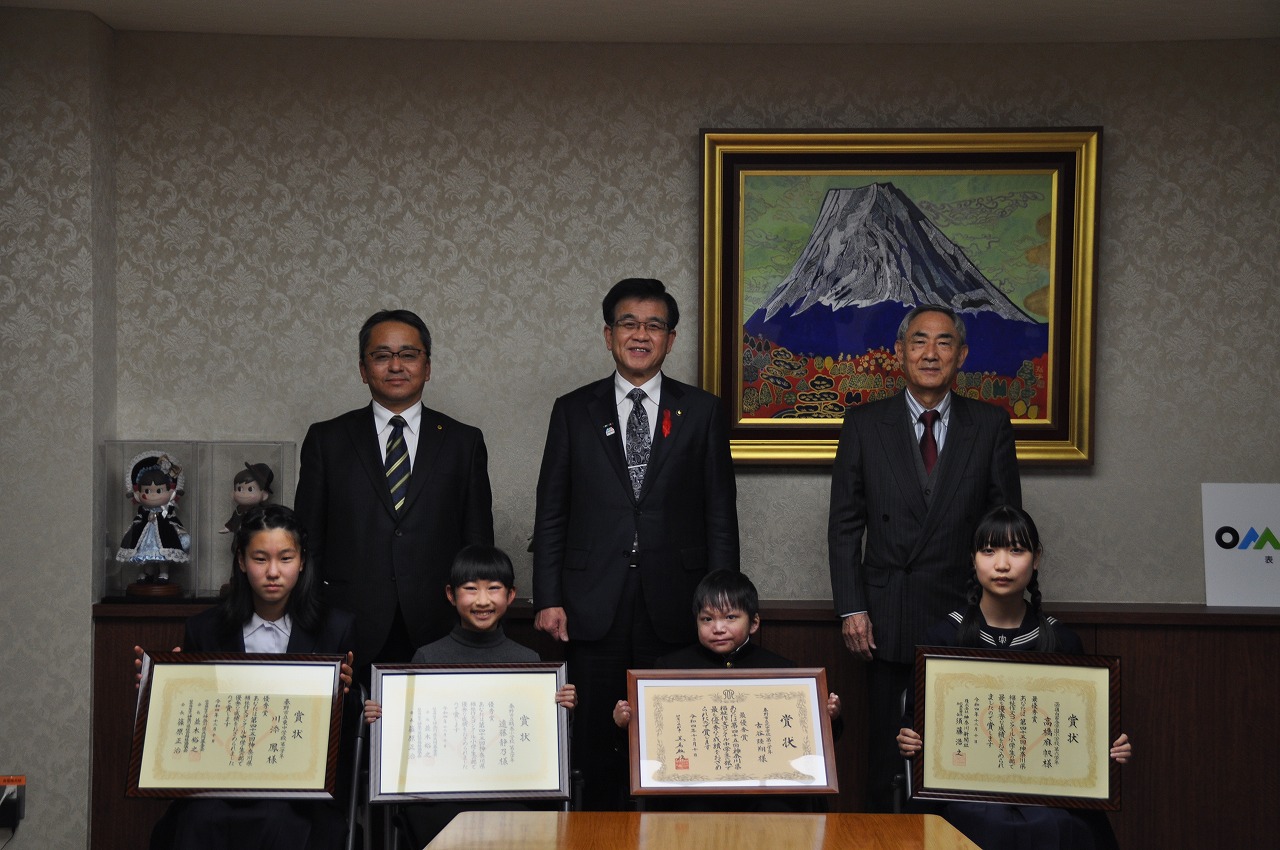 応接室で市長を含めた3人の男性と四人の子どもが並んでおり、子どもたちはそれぞれ額に入った表彰状を手にしている写真