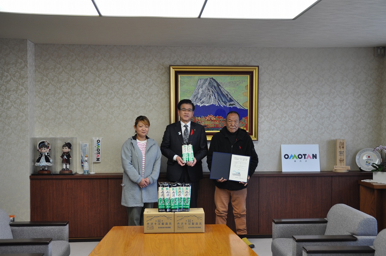 応接室で市長を挟んで、左右に男女が立ち、市長が年越しそばが包装された袋を持ち、右の男性は賞状とみられる額を抱えている写真