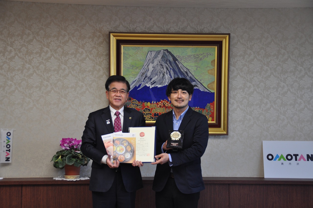 応接室でココハダLAB・ジェントルゆうすけ代表と市長が並んでおり、左の市長は雑誌と賞状を持ち、右のゆうすけ代表はトロフィーを持って笑顔でカメラを見つめている写真