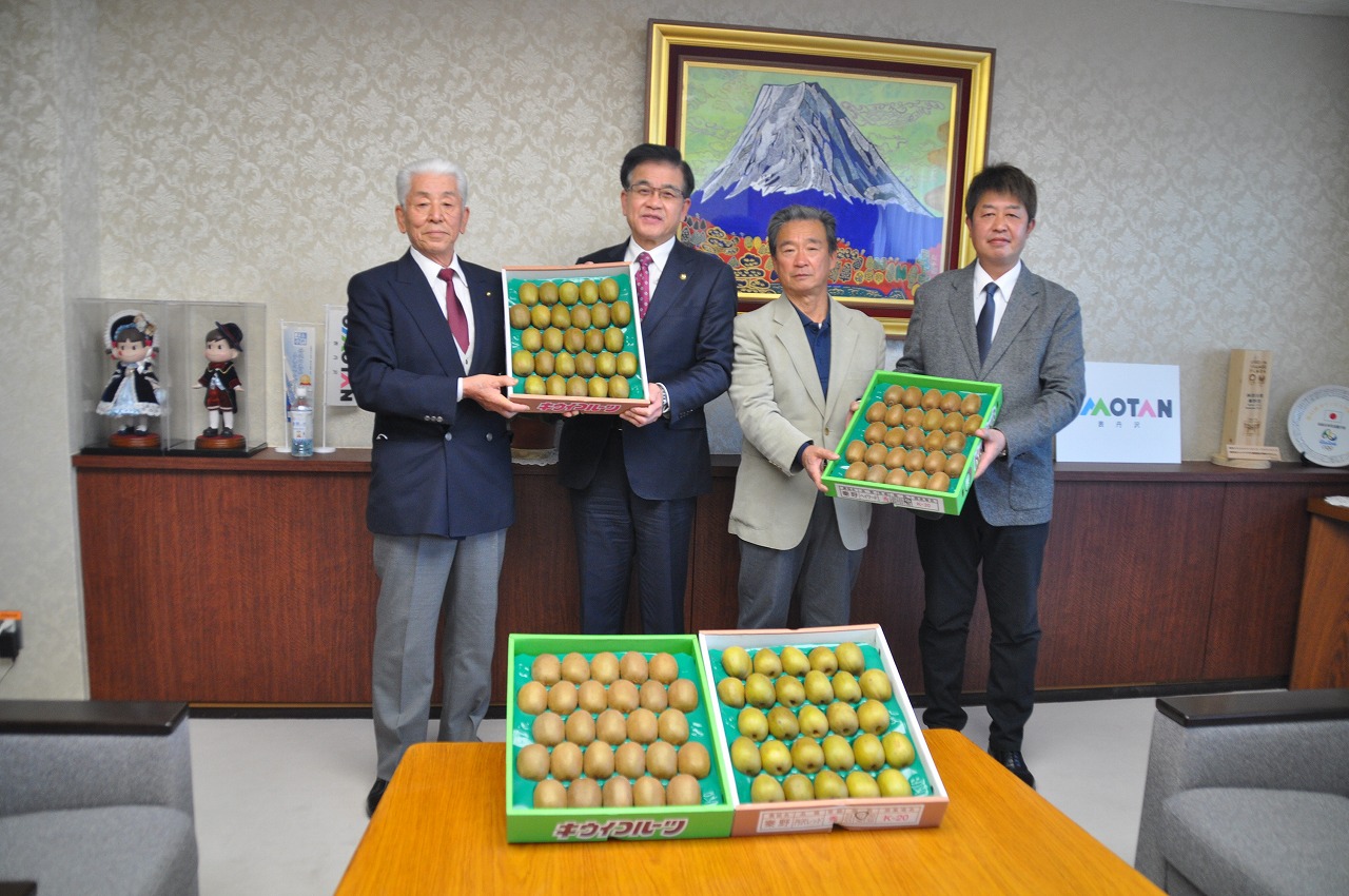 応接室で市長を含めたスーツ姿の男性四人が並んでおり、机と両手で抱えている計4つの箱に入った大量のキウイフルーツをカメラに向けて記念撮影を行っている写真