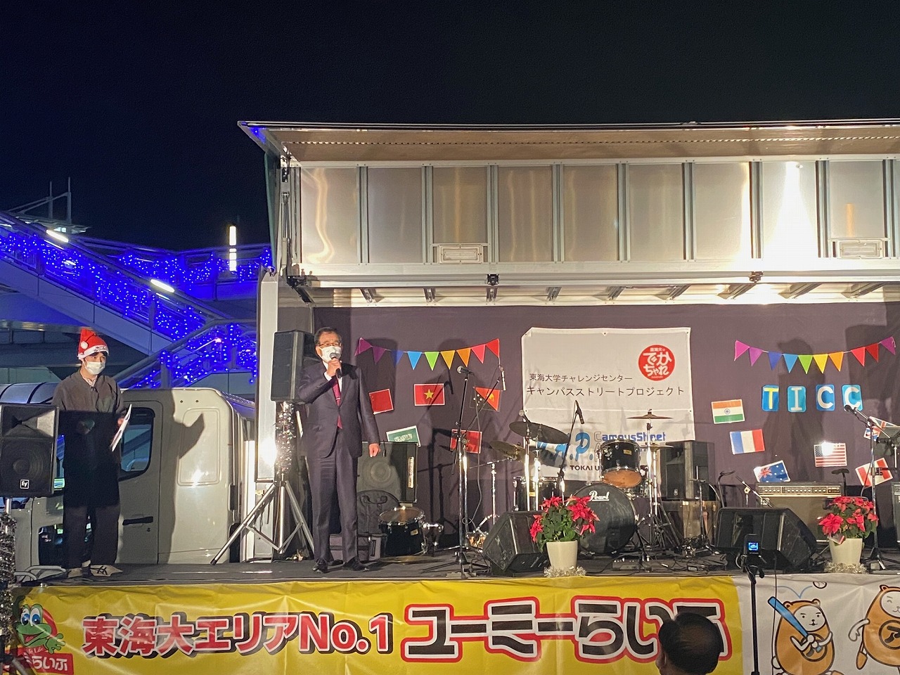 野外ステージに立つ二人の男性と、その後ろに楽器が置かれており、ステージ上には世界各国の国旗を模した旗が飾られている夜のイベント会場の写真