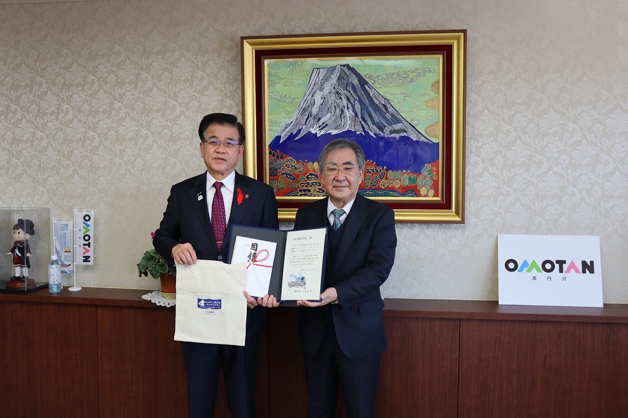 応接室で市長とスーツ姿の男性二人が並んでおり、目録とみられるものを持ってカメラを見つめている写真