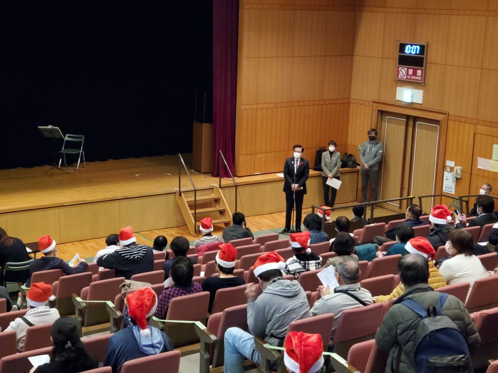 広々としたホールで、サンタクロースの帽子をかぶった観客たちが客席に座っており、ステージ下に設置されたマイクの前で男性がスピーチをしている様子の写真