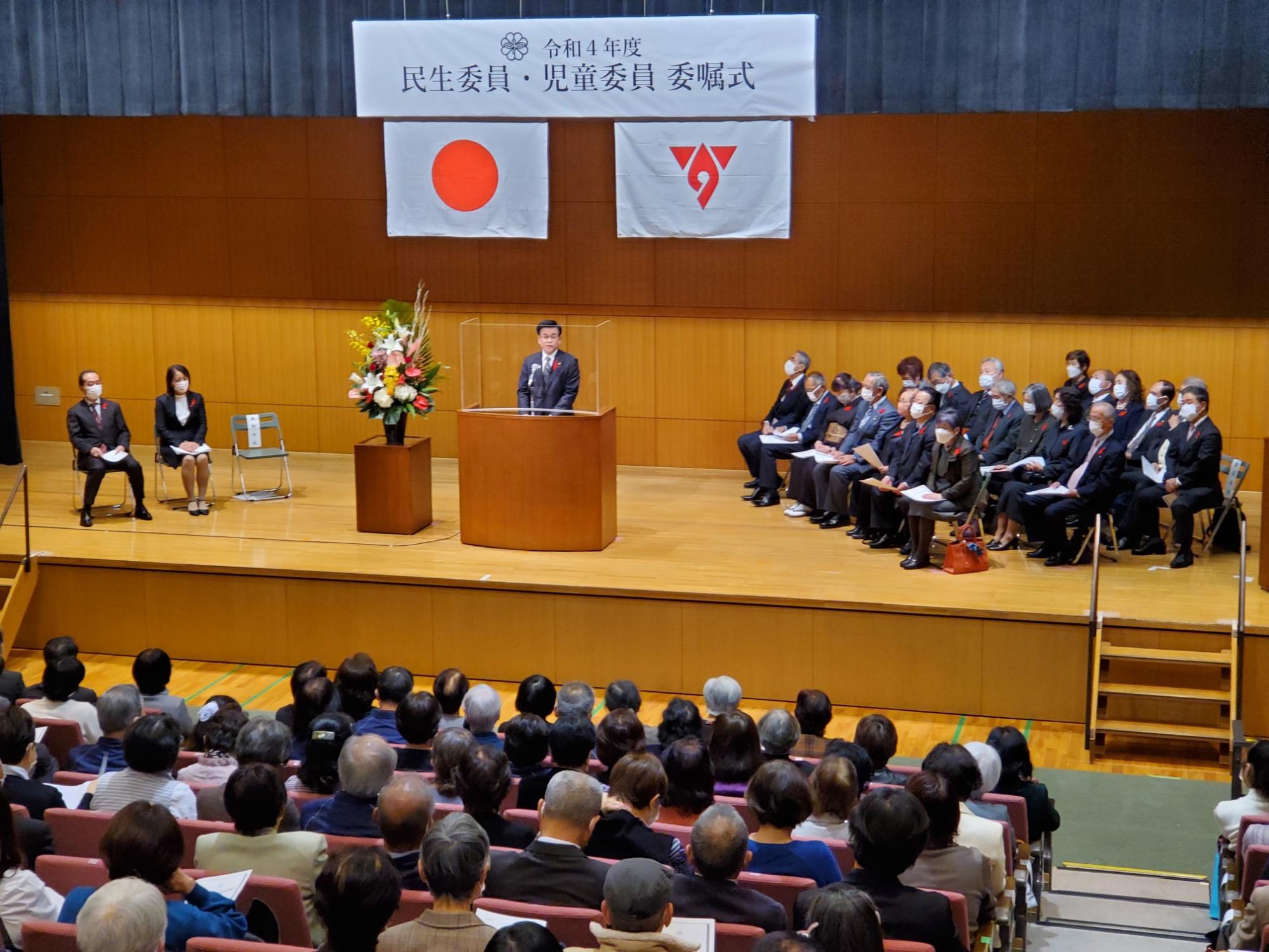「民生委員・児童委員委嘱式」の横断幕が掲げられている式典が行われている講堂で、壇上では市長がマイクを使ってスピーチし、座席には多くの人が座っている写真