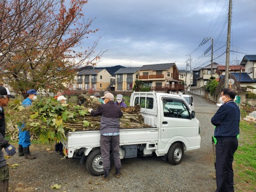 空き地のような場所で、数人の人々が軽トラックの荷台に剪定された木の枝を積んでいる様子の写真