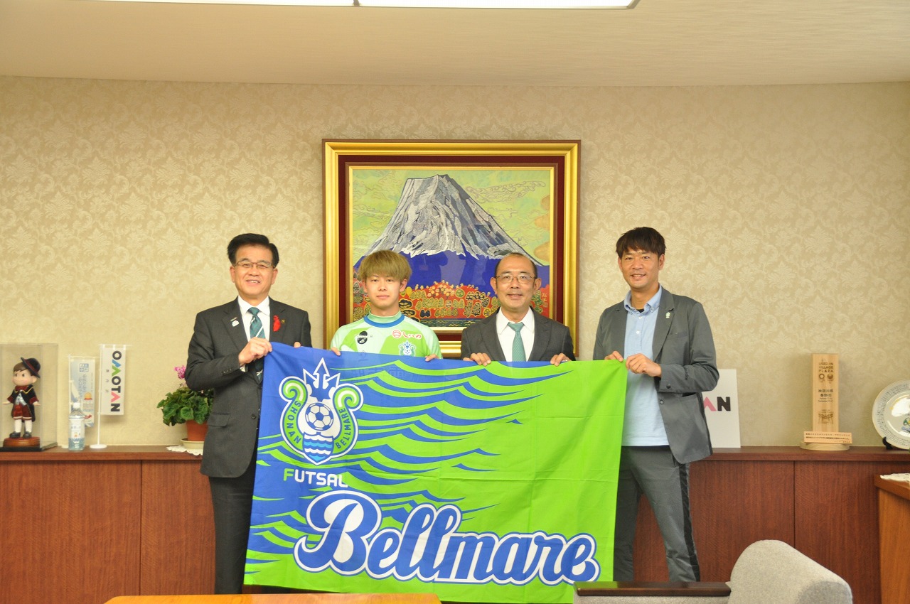 応接室でスーツ姿の男性三人とユニフォームを着た男性一人が、湘南ベルマーレフットサルチームのロゴが入った大きな緑と青の旗を広げている写真