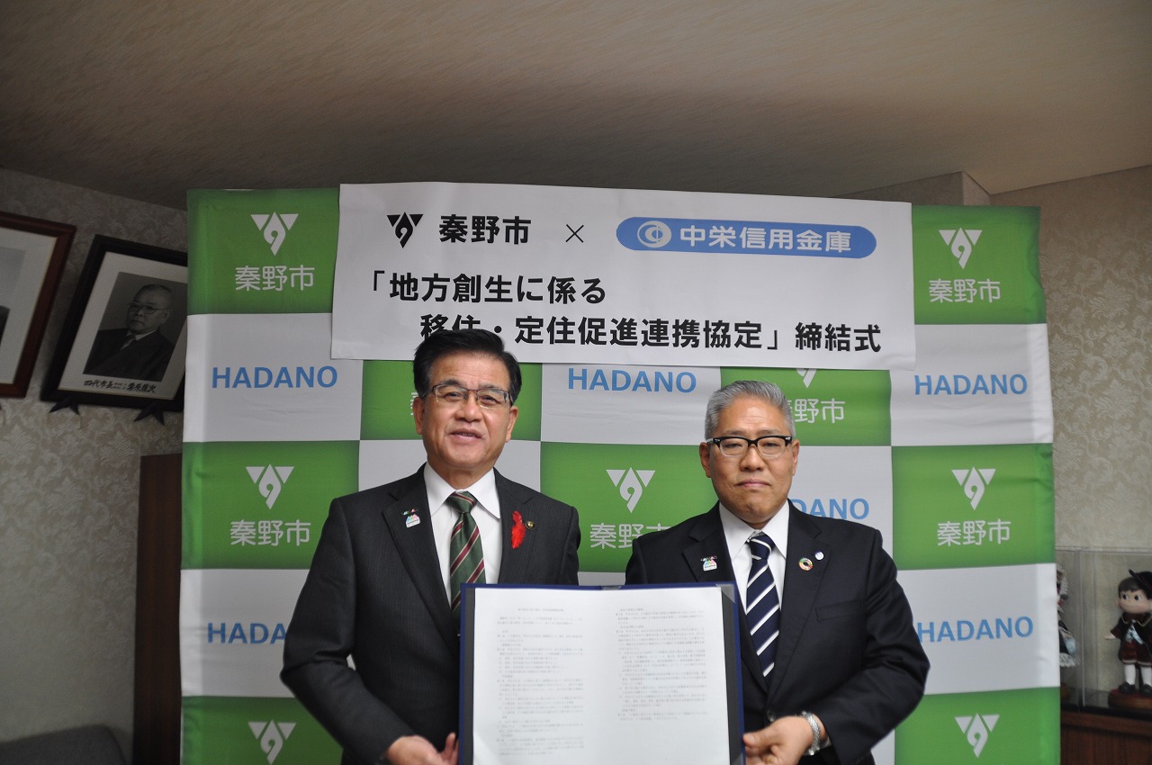 記者会見場で、市長とスーツ姿の男性二人が「地方創生に係る移住・定住促進連携協定締結式」と書かれた看板の前で、署名が入った書面を持ってカメラに笑顔を向けている写真