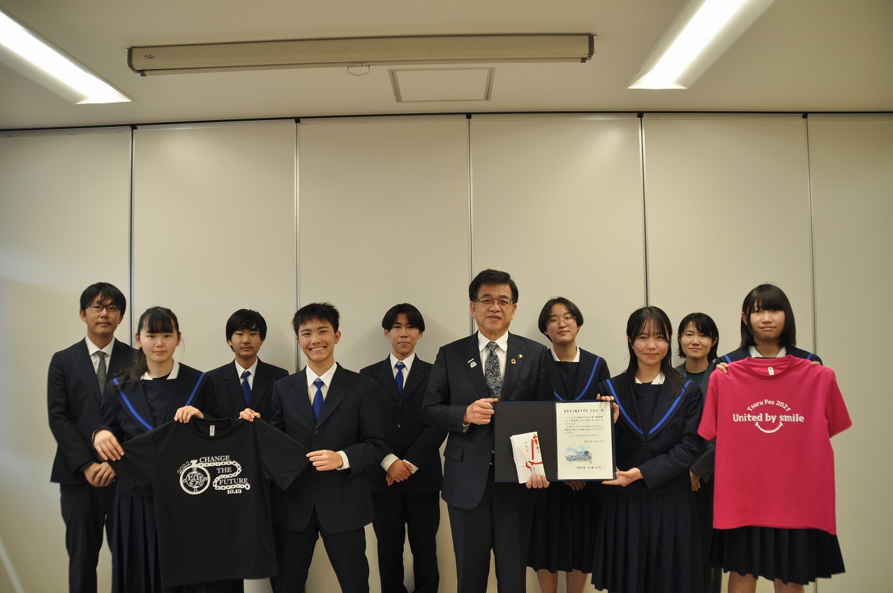 スーツ姿の市長を中心に、制服を着た生徒たちが並び、目録や黒とピンクのTシャツを持って笑顔で記念撮影している写真