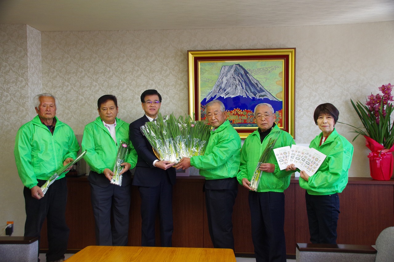 緑のジャンパーを着たJAはだの葉ニンニク研究会の5人の方々とスーツ姿の市長が並び、たくさんの葉ニンニクが入ったかごやレシピ集を手にして記念撮影している写真