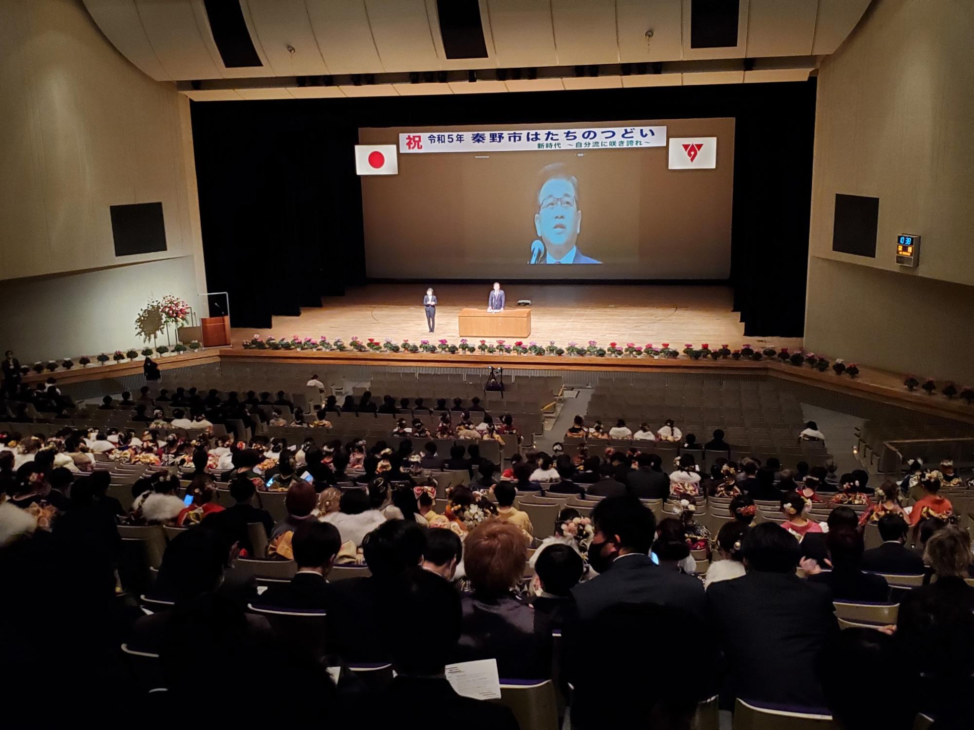 大きなホールの観客席にスーツや晴れ着姿のたくさんの出席者が着席し、舞台上の演台で挨拶する市長が大型スクリーンに映し出されているはたちのつどいの様子を後方から写した写真