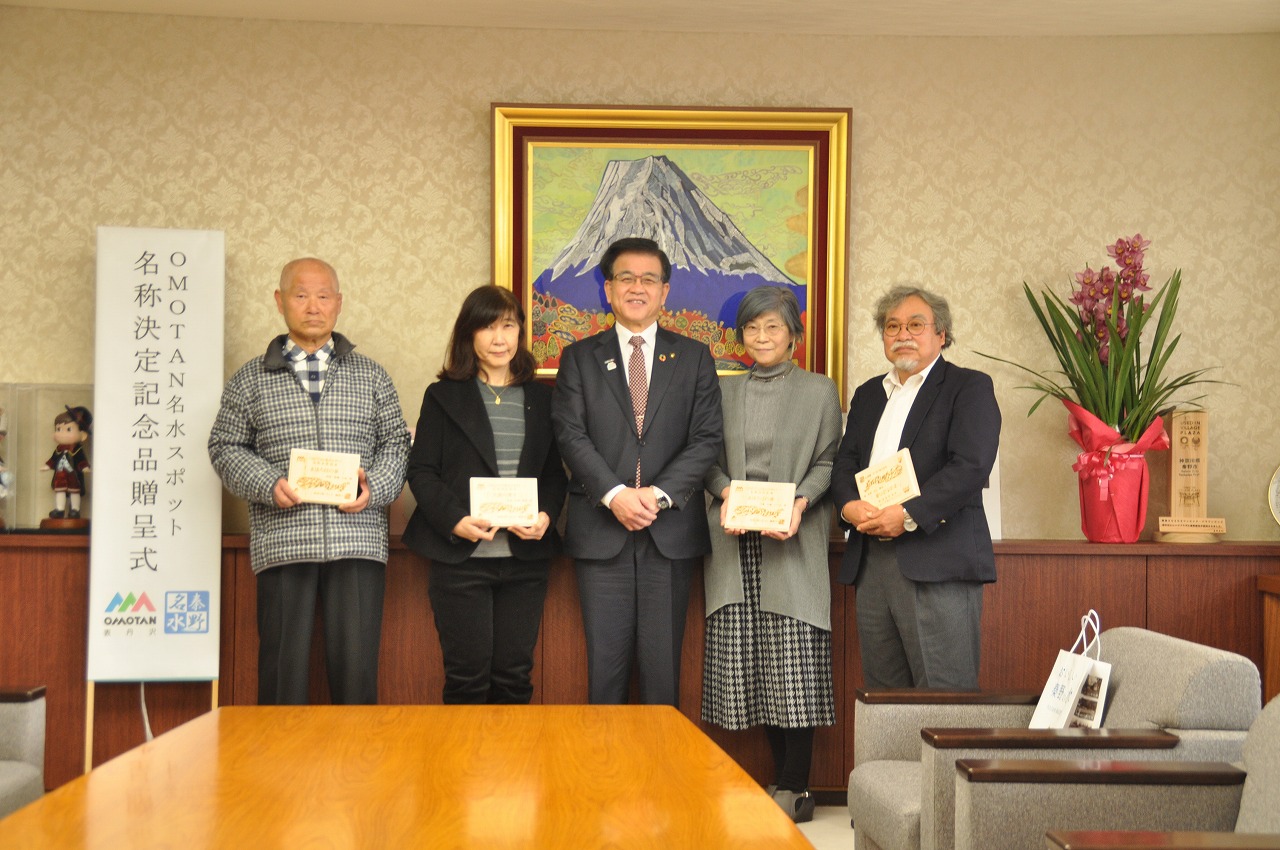 「OMOTAN名水スポット名称決定記念品贈呈式」の立て看板の横で、スーツ姿の市長を中央に、4人方々が木製の記念品を手に並んで立っている記念写真