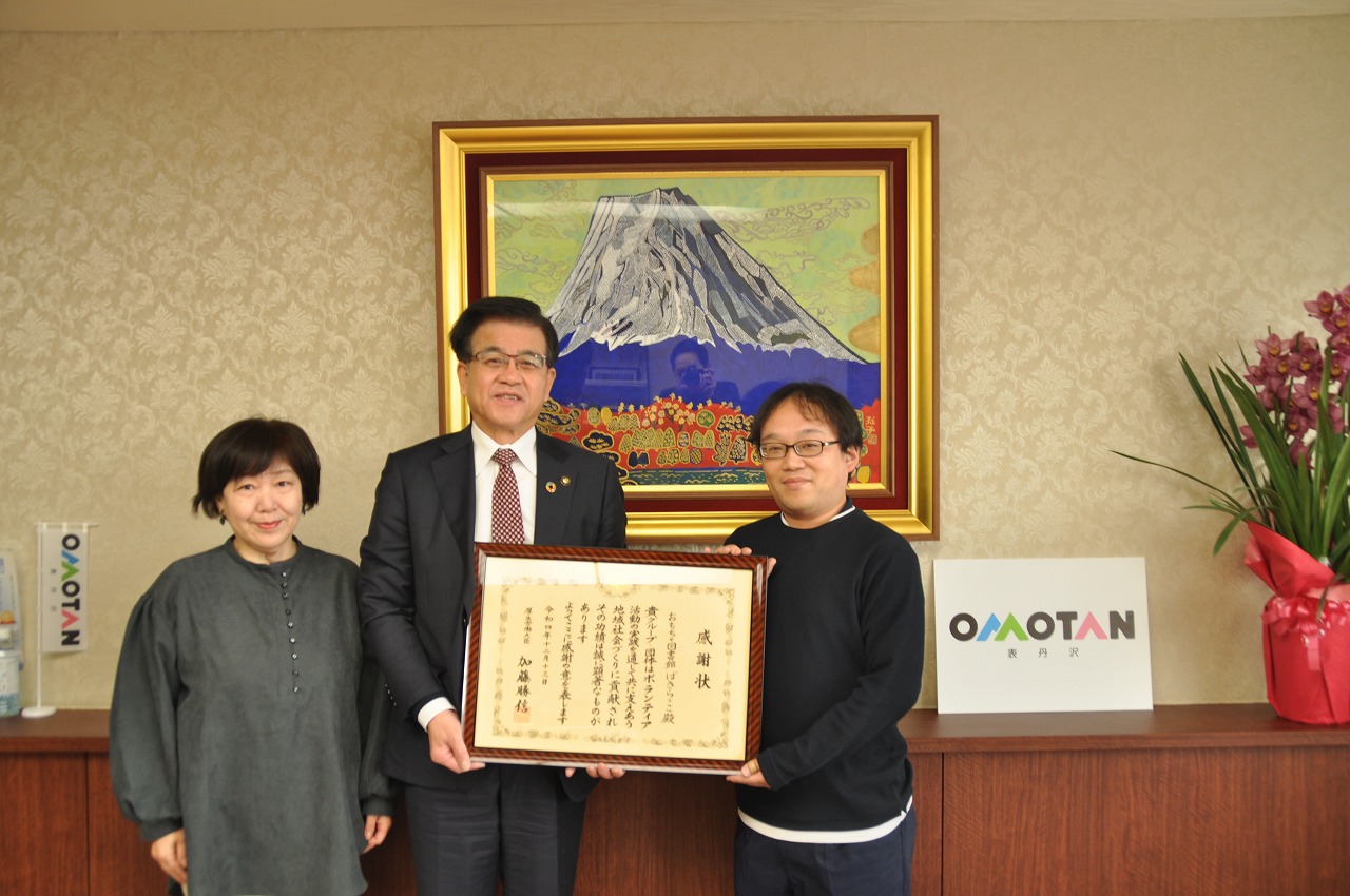 スーツ姿の市長と山の絵画が掛かった壁の前で、市長と眼鏡をかけた男性が大きな額に入った感謝状を左右から持ち、市長の隣に並んだ女性と一緒に写した記念写真