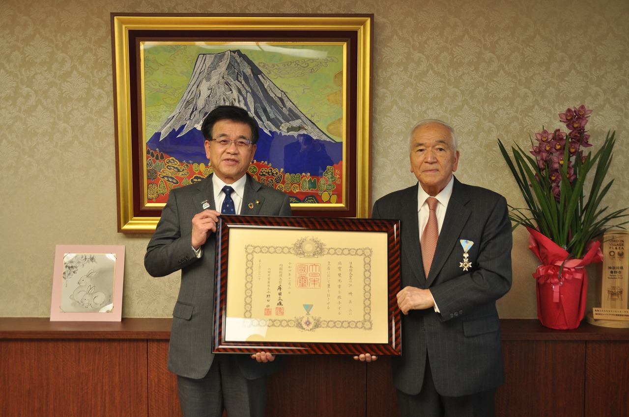 市長とスーツの上着に胸に勲章を着けた男性が並んで立ち、大きな額縁入りの表彰状を一緒に持ち記念撮影している写真