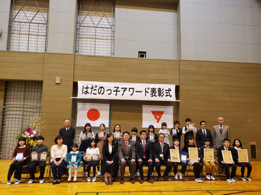 「はだのっ子アワード表彰式」の横断幕と日本国旗と市章の旗が掲げられた体育館の壁の前で、市長を中央に、賞状や盾を手にした子どもたちと関係者が整列し、前列は椅子に座り、後列は立って写した記念写真