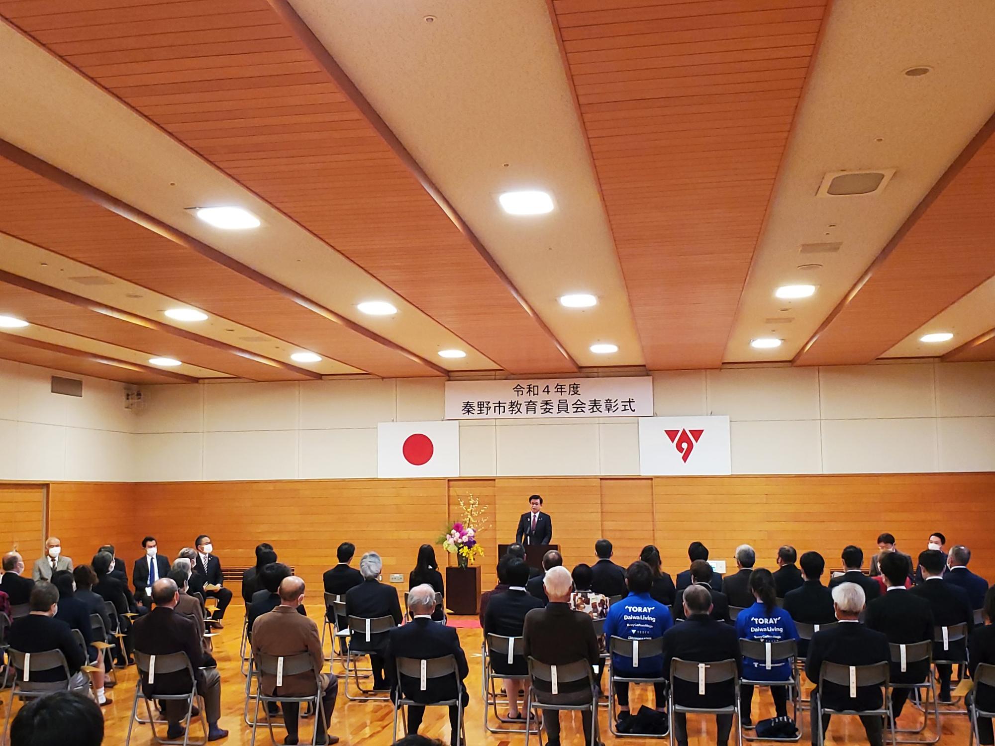 「令和4年度 秦野市教育委員会表彰式」の横断幕と日本国旗と市章が掲げられた壁の前に置かれた演台で市長が話をしており、前方に出席者たちが整然と並んだ椅子に着席している表彰式の写真