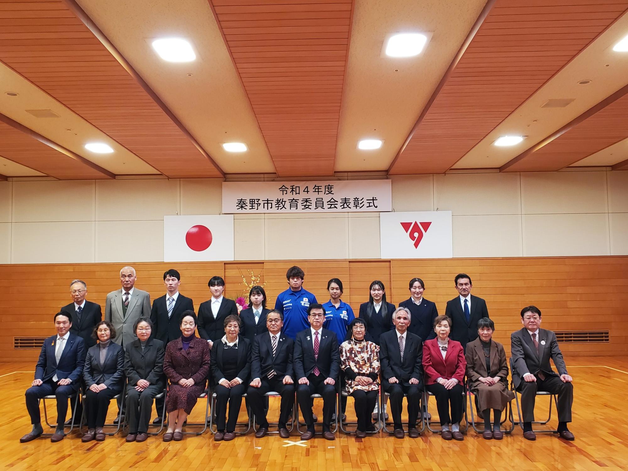 「令和4年度 秦野市教育委員会表彰式」の横断幕と日本国旗と市章が掲げられた壁の前で、市長を中央にしてたくさんの関係者が集まり、前列は椅子に座り、後列は立って写した記念写真