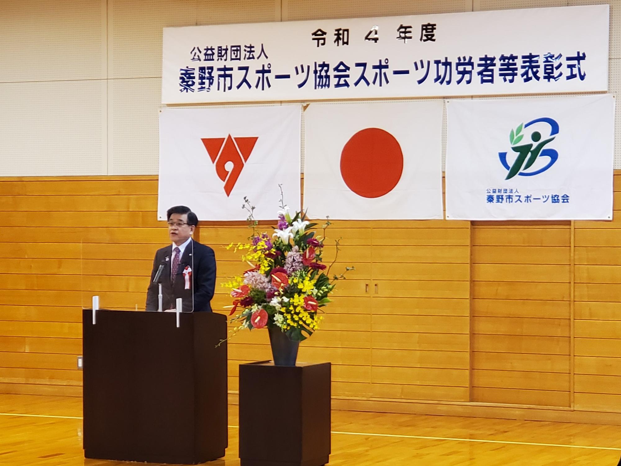 式典会場の演台に立つ市長がスピーチをしている様子を、背景の壁に掲げられた秦野市スポーツ功労者等表彰式の横断幕や旗と共に捉えた写真