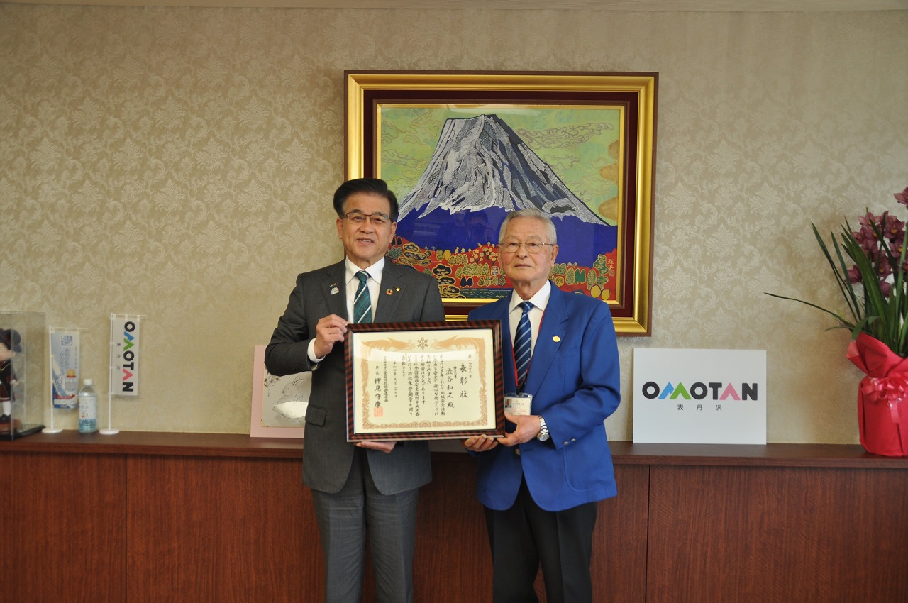 スーツ姿の市長が青いジャケットの男性とともに、額に入った表彰状を手にカメラに向かって微笑んでいる様子を捉えた写真