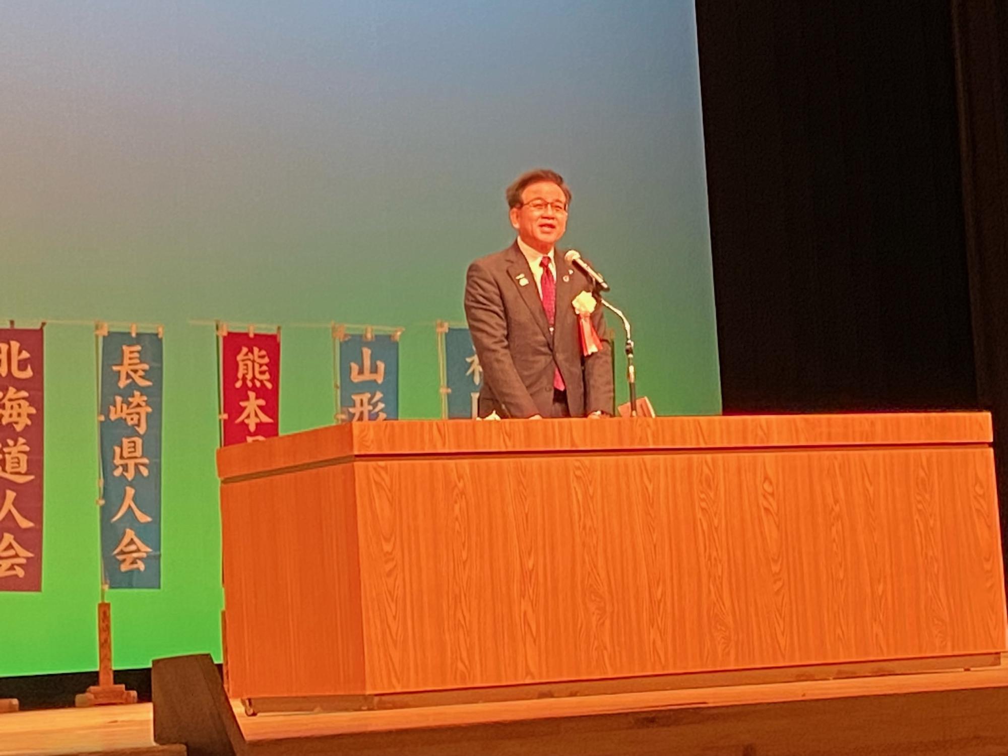 檀上に設置された木製の演台で、スーツを着た市長がマイクに向かってスピーチをしている様子を、背景に並んだ「北海道人会」「長崎県人会」などののぼり旗と共に捉えた写真