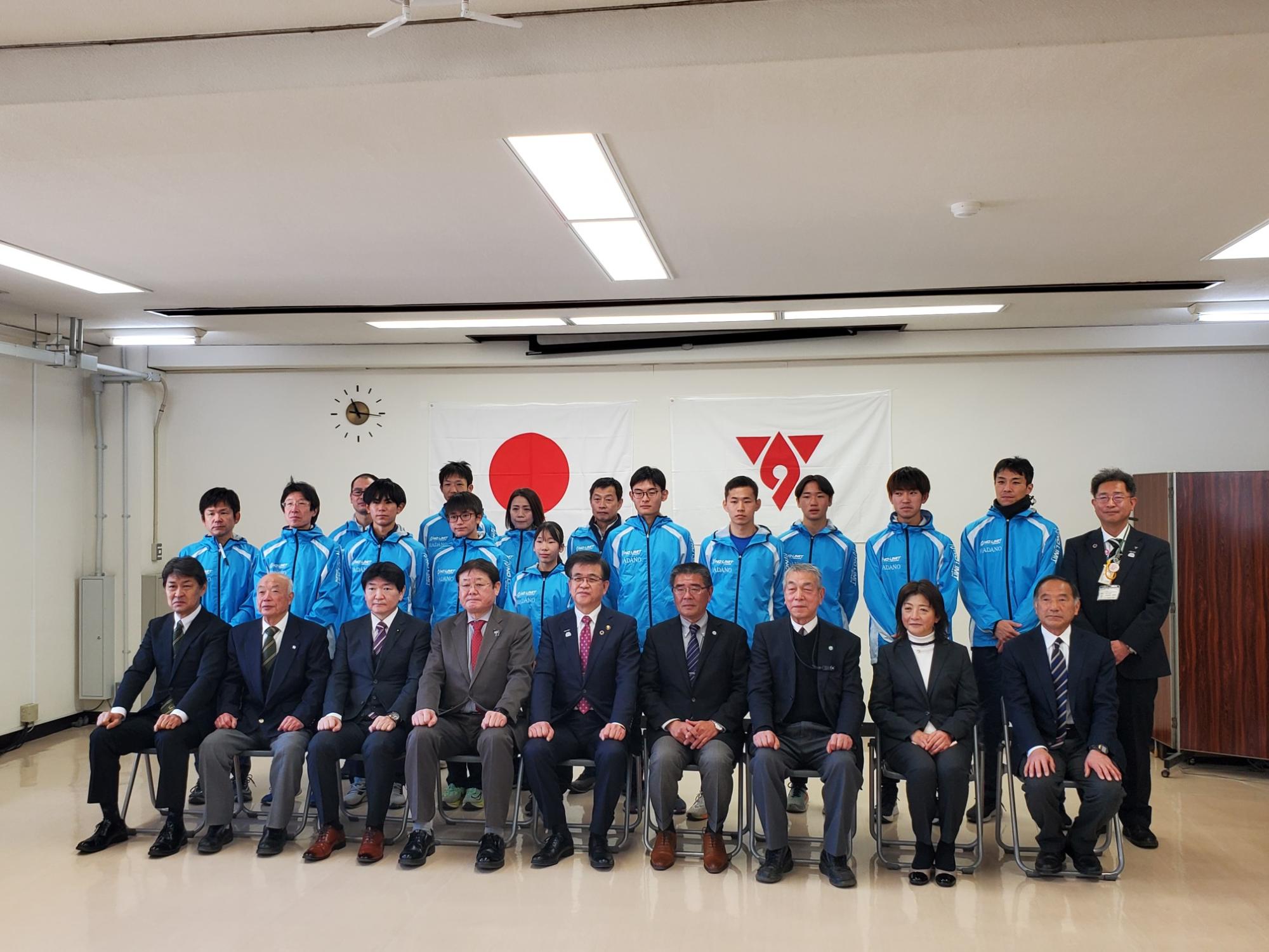 背景に日の丸とロゴの旗が飾られた部屋で、スーツ姿の市長と男性たちと、青いジャージを着た若者たちが、二列になって集合記念撮影をしている様子を捉えた写真