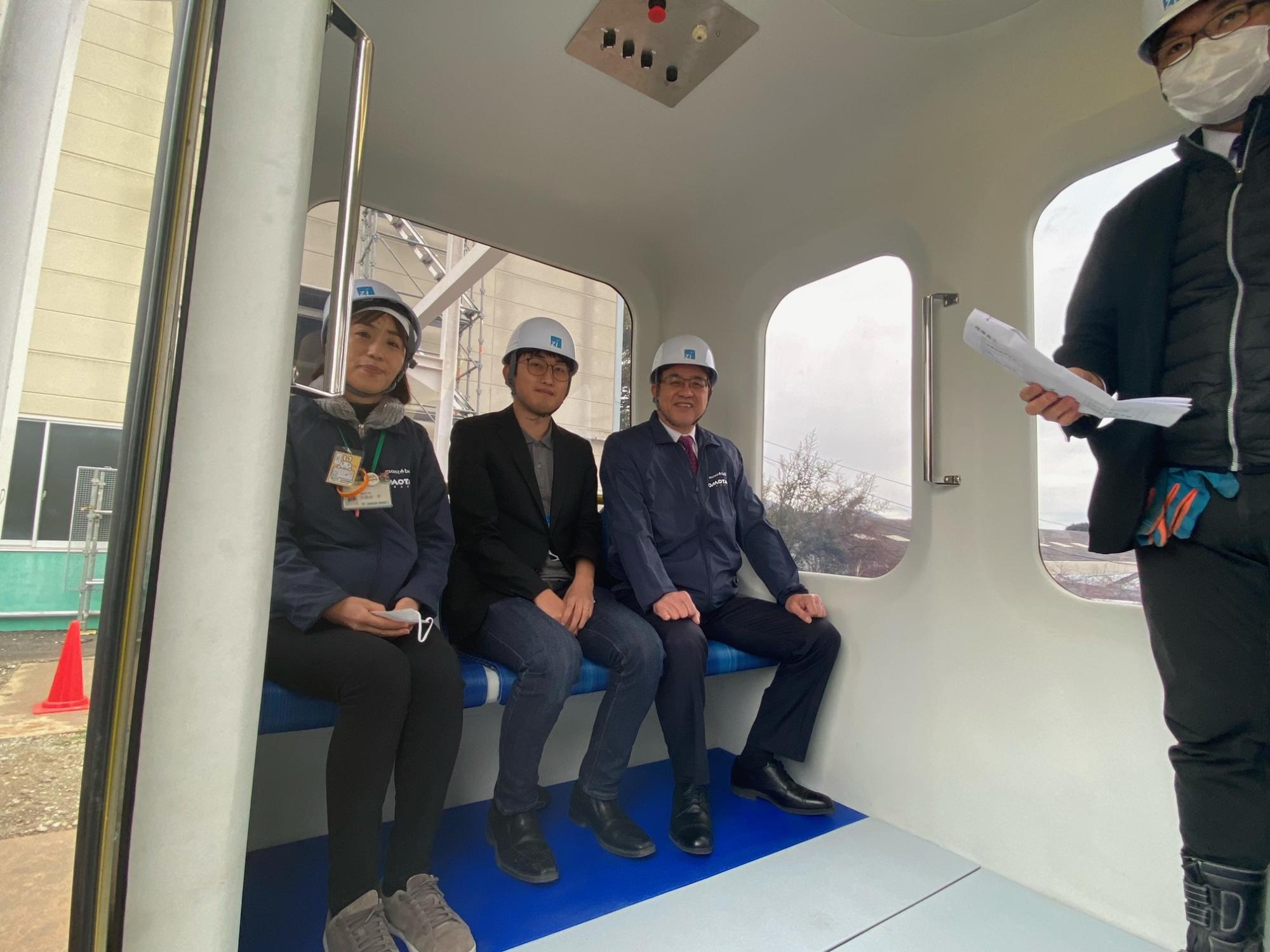 工事用のヘルメットを被った複数の男女が、新しい交通システムの小型車両の内部に座って、その乗り心地などを確認している様子を捉えた写真